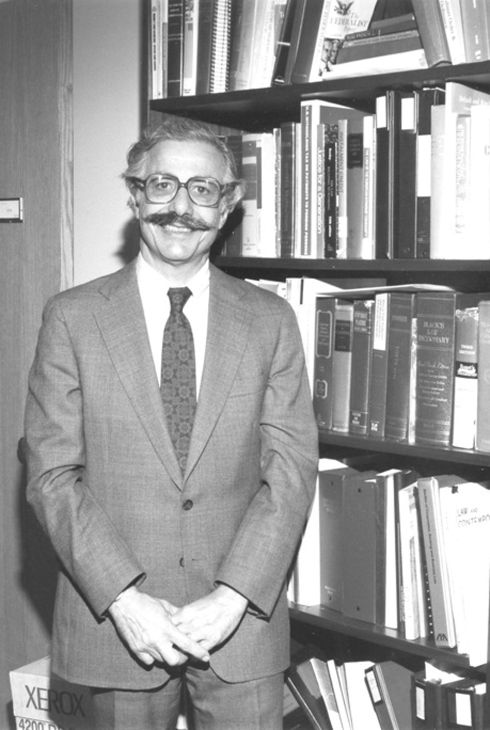 black and white image of Carl Felsenfeld standing in an office, next to a packed book case