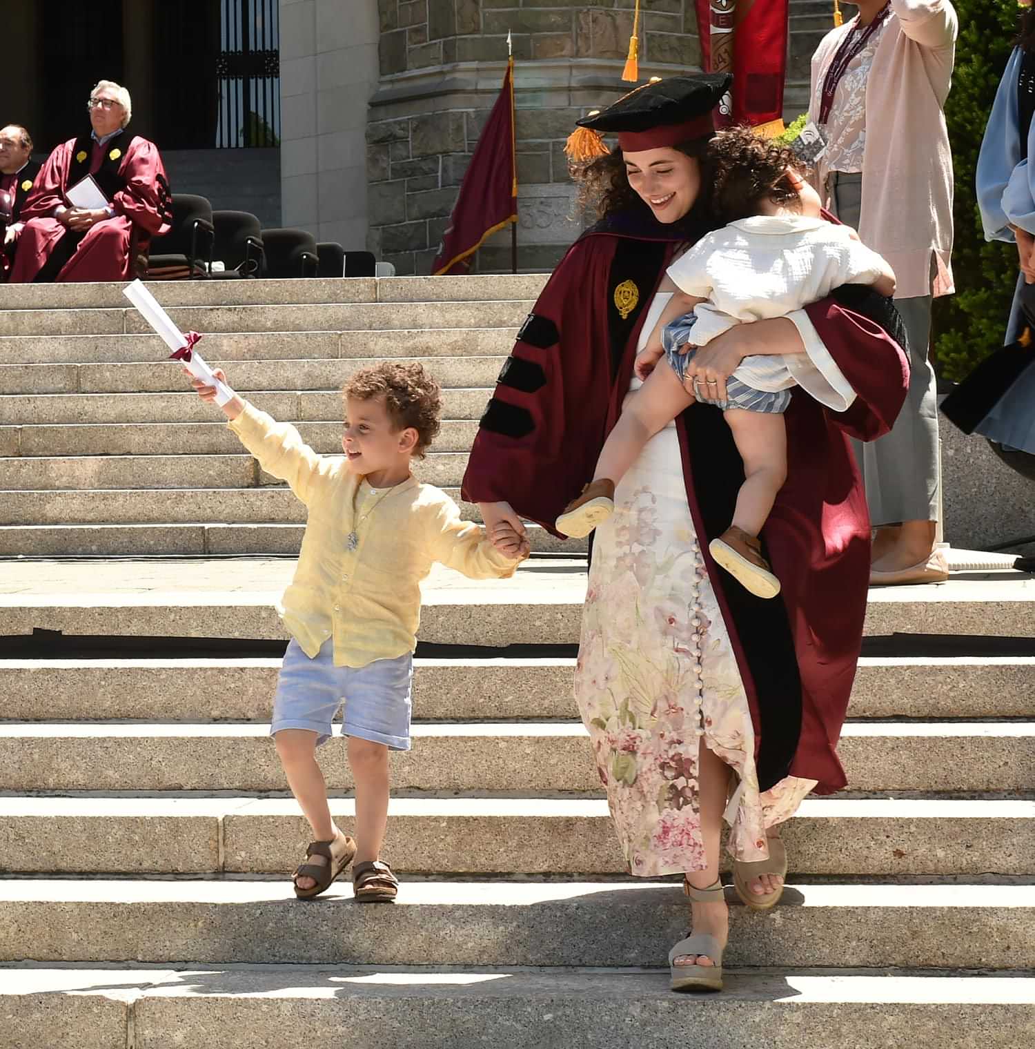 a graduate holds a sleeping baby and the hand of a toddler after receiving a diploma on stage