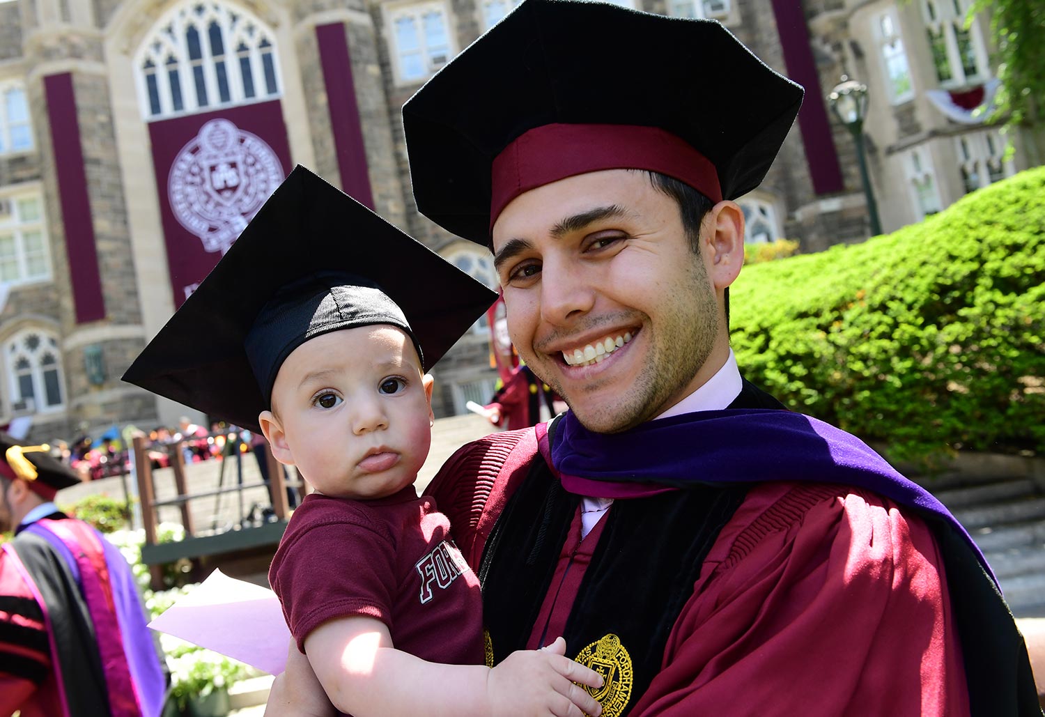 a smiling graduate holds a baby, both wearing caps