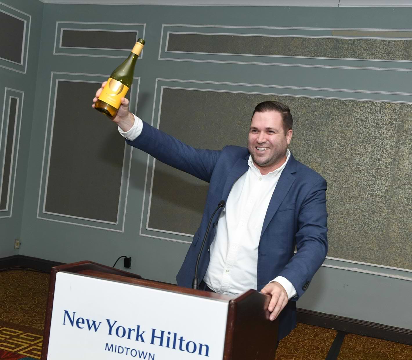 a alumni stands a podium holding a bottle of wine
