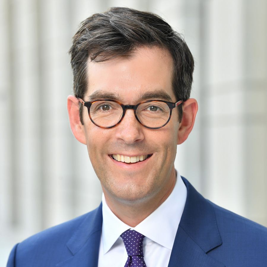 John R. Brooks smiles for a headshot wearing thick rimmed round glasses, a blue suit and a deep purple and white polka dot tie