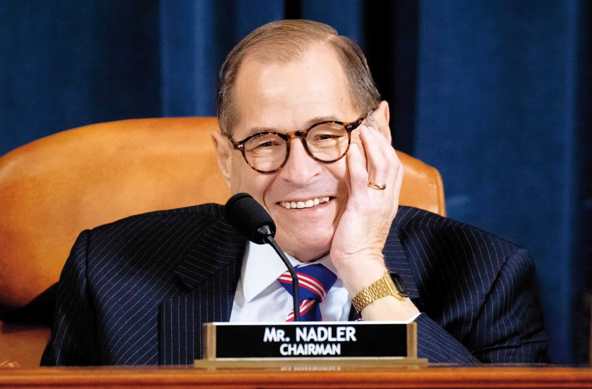 close view of Jerry Nadler smiling while sitting at mircophoned table, a name plate resting in front of him reads "Mr. Nadler, Chairman"