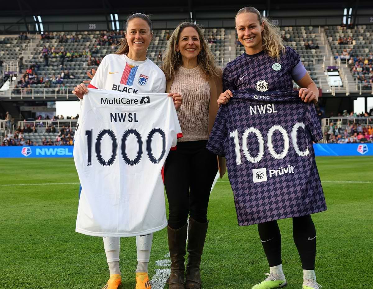 Jessica Berman smiles while standing on a soccer pitch between two NWSL players holding jerseys with the number 1000