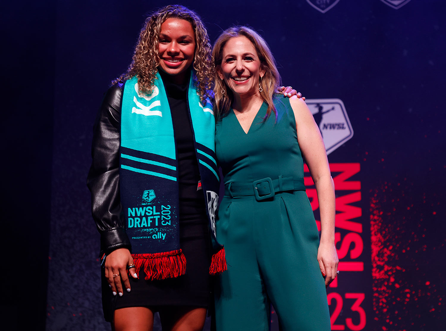 Jessica Berman smiles while standing on the NWSL 2023 Draft Pick stage beside a woman soccer athlete