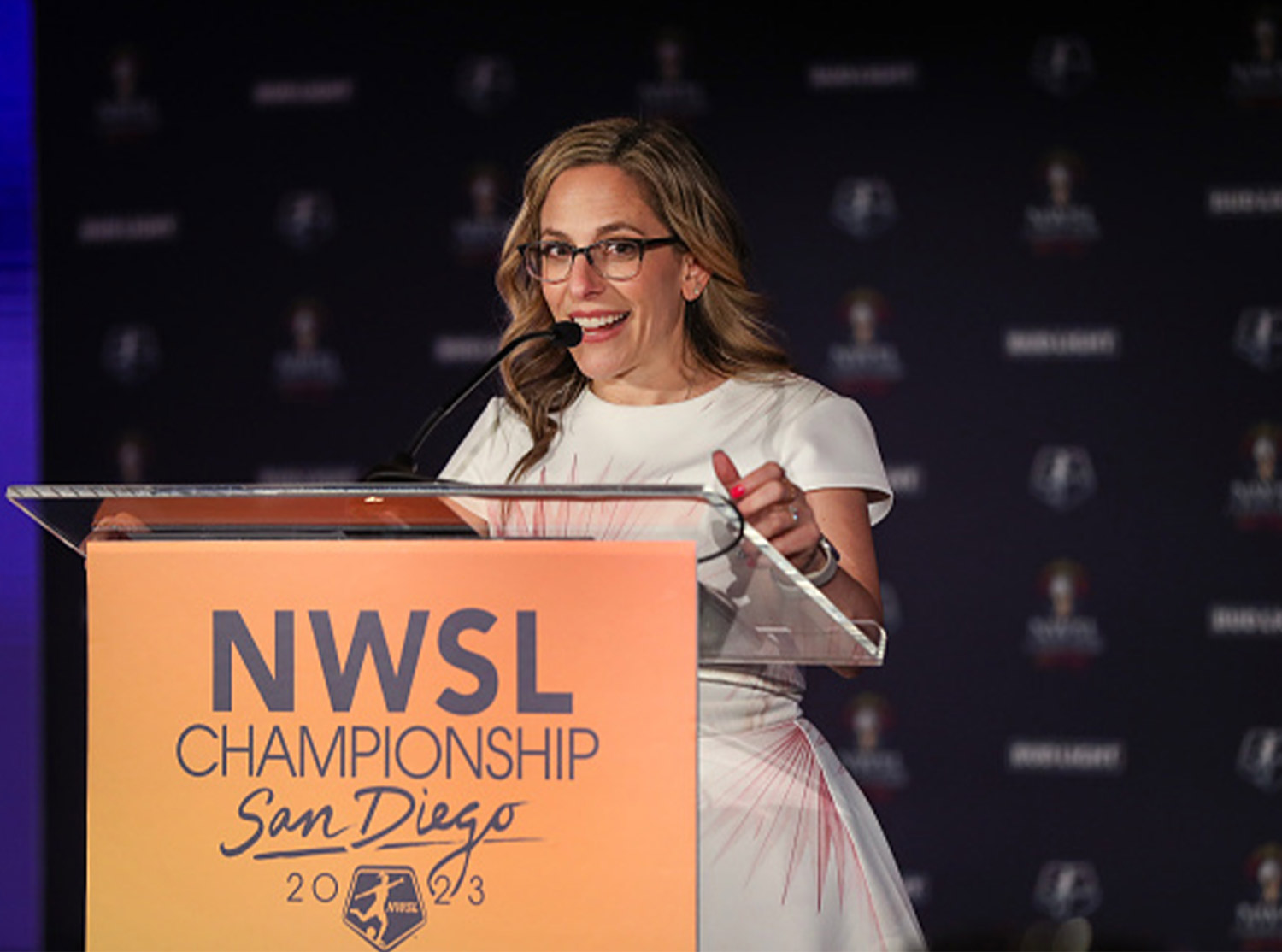 Jessica Berman wears glasses and a white dress with pink accents as she speaks at the podium of the NWSL Championship 2023 in San Diego