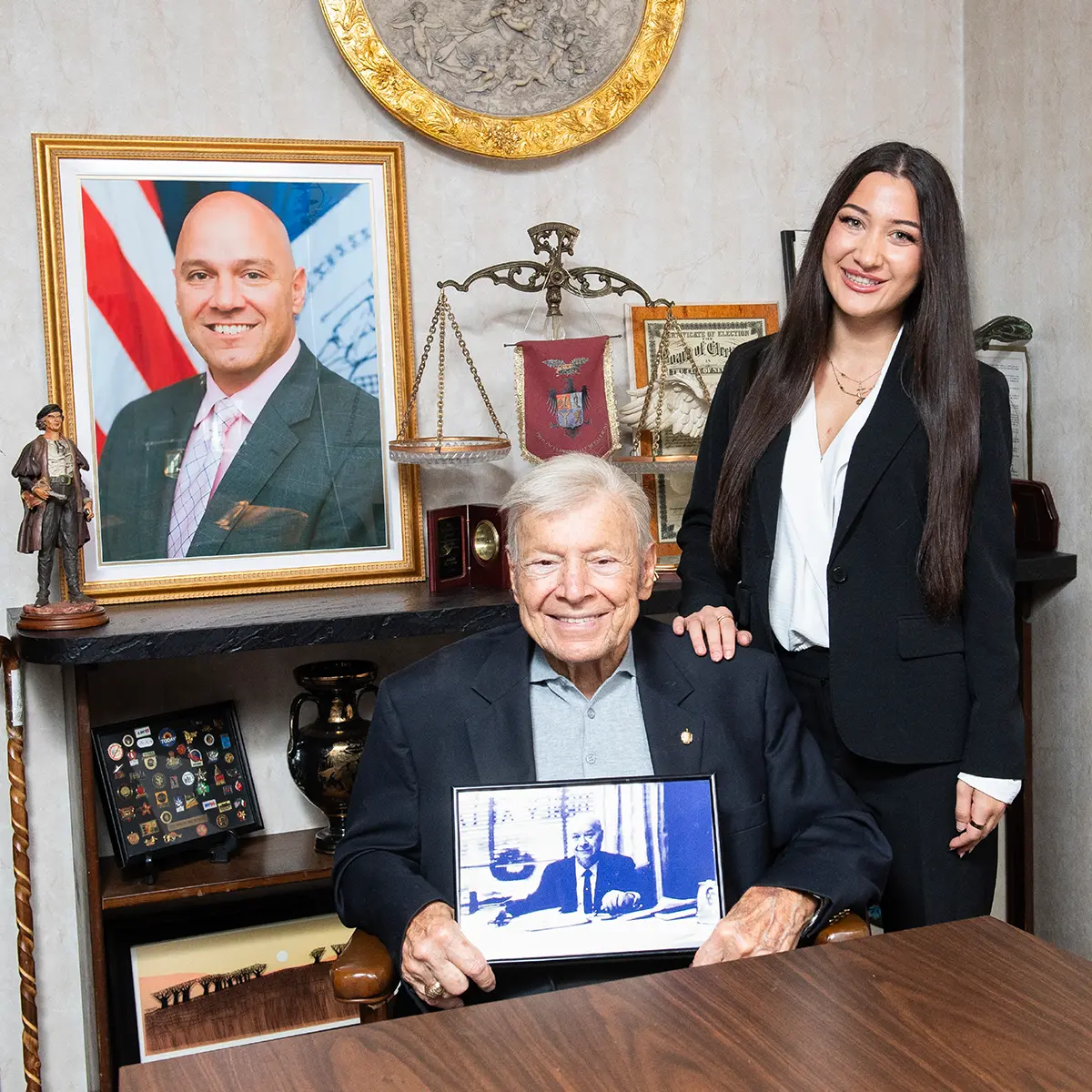 In an office, an older man in a suit jacket sits at a desk holding a framed photo, while a younger woman in a black suit stands behind him. The background features a framed portrait of another man, a scales of justice emblem, and various other framed documents and decorative objects on shelves and the wall.