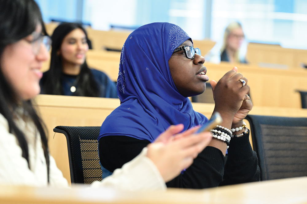 In a tiered classroom, a student wearing a bright blue, embellished hijab and glasses gestures with her hands while looking towards the right. Two other students are visible in the foreground and background, also appearing engaged in the classroom activity.