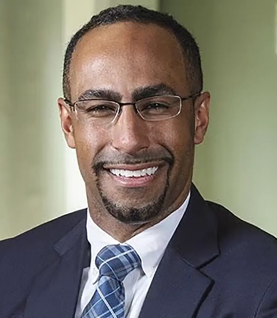 Portrait orientation photograph headshot of Jerry Dickinson, a smiling bearded man wearing a dark navy blue business blazer suit and a white button-up dress shirt underneath with a multi-colored pattern tie equipped (marine blue/white)