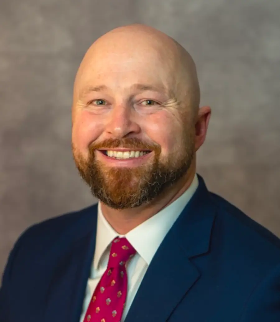 Portrait orientation photograph headshot of Prescott Loveland, a smiling bearded man in a dark navy blue business blazer suit and white button-up dress shirt underneath with a multi-colored pattern tie (hot dark pink/chrome)