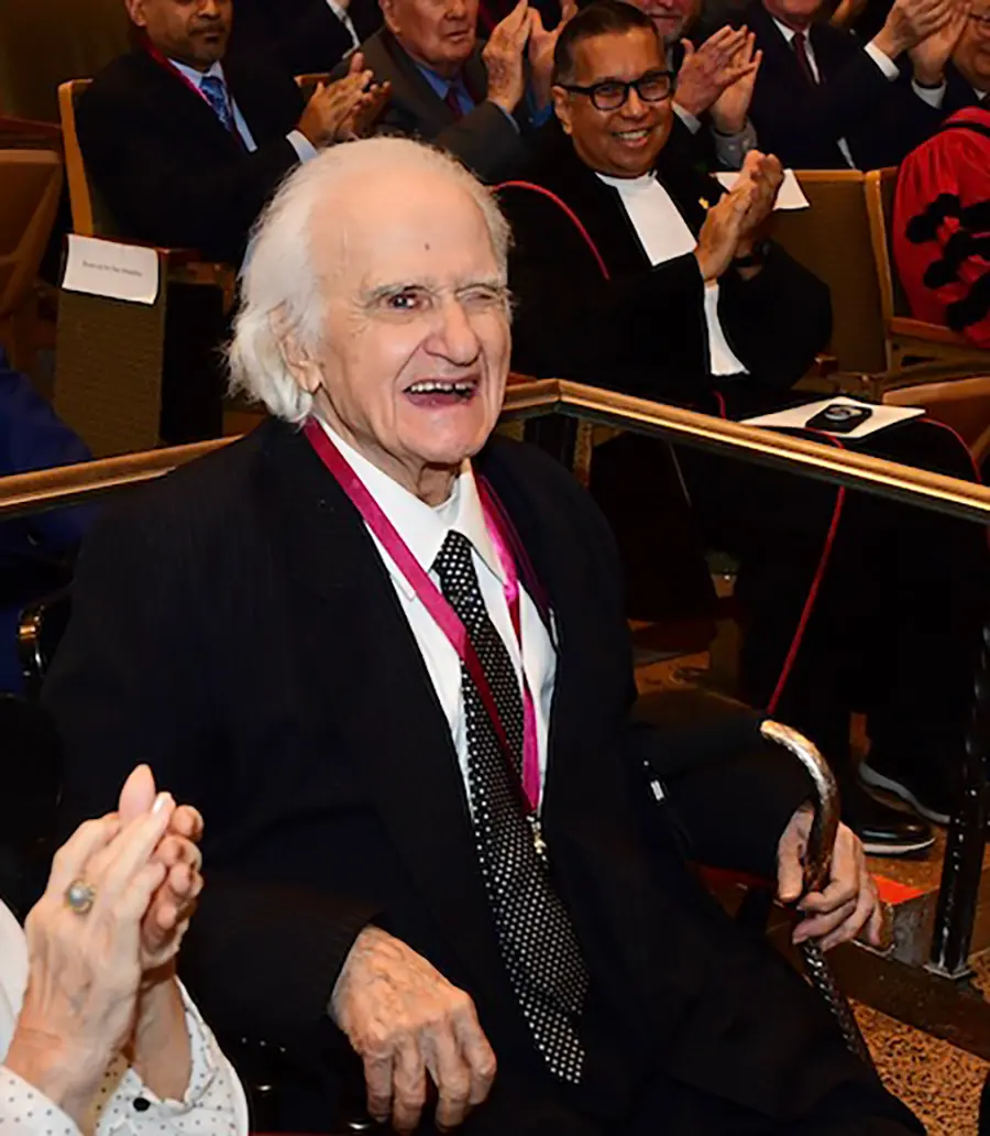 Portrait orientation photograph headshot of Professor Constantine N. Katsoris , a smiling man in a black business blazer suit and white button-up dress shirt underneath with a multi-colored pattern tie (black/white) as he is seated down in an auditorium chair setting with other people around him seated down smiling as they clap their hands