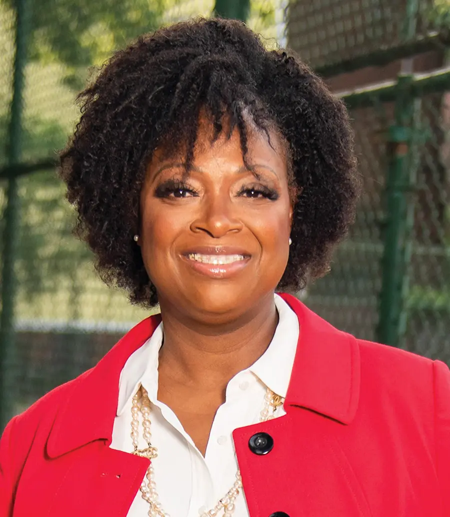 Portrait orientation outdoor photograph headshot of Valerie White, a smiling woman in a dark red coat and white button-up dress shirt underneath with off-white pearl necklaces
