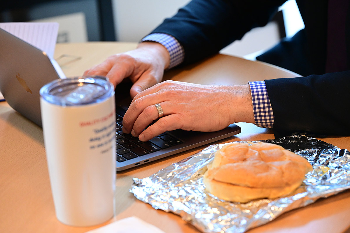 A person's hands type on a laptop keyboard next to a sandwich on foil and a white tumbler.