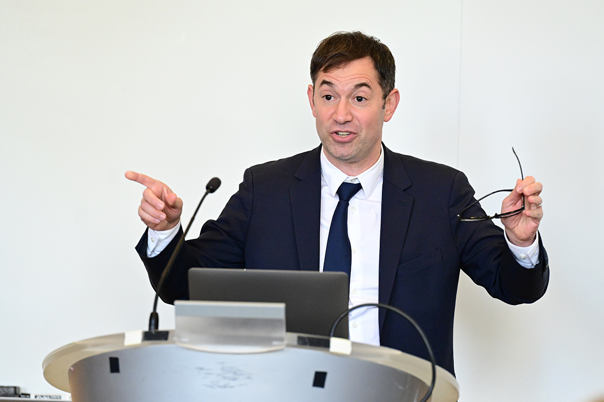 Joseph Landau wearing a suit stands at a podium, gesturing and holding glasses.