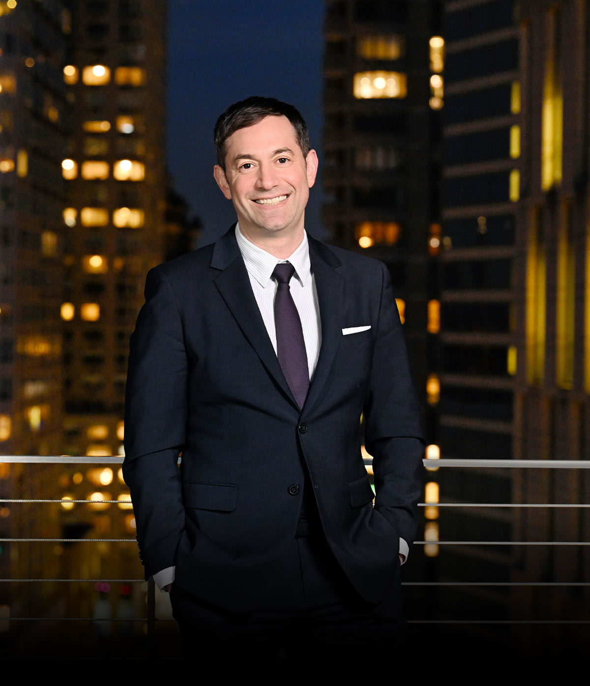 Joseph Landau wearing a dark suit stands on an outdoor balcony at night, with city buildings and illuminated windows in the background.