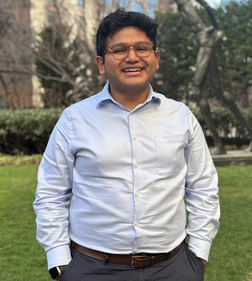 A smiling, portrait image of Javier Melendez, a male student with dark hair and glasses. He is standing outdoors on a patch of grass with a building in the background, wearing a light blue button-down shirt and dark trousers with a brown belt.