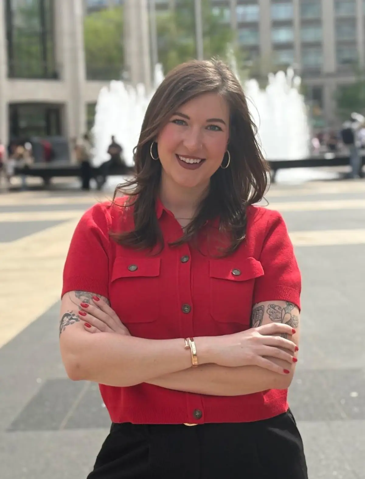Portrait orientation outdoor photograph of Megan McWaters, a smiling woman with dark brown colored hair and tattooed arms standing outside with her arms crossed; She is wearing a red, short sleeved button-up dress top and black pants; A fountain, buildings, and people are visible in the blurred background of a plaza behind her