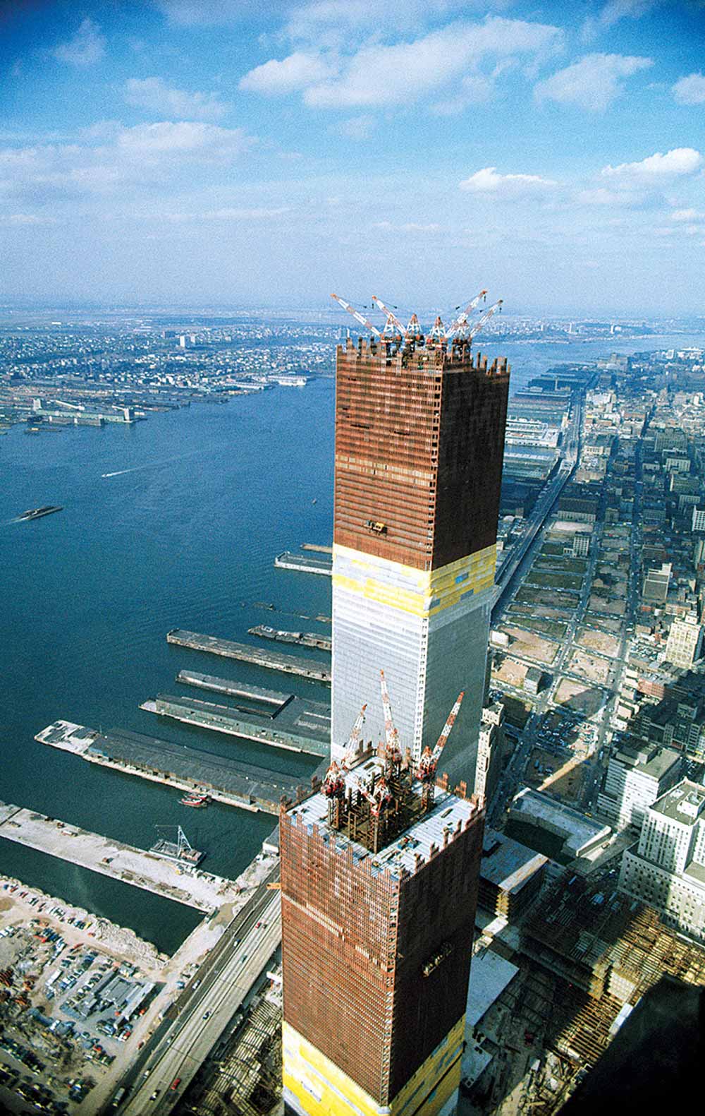 An aerial view of the original World Trade Center towers under construction in New York City, overlooking the Hudson River and piers.