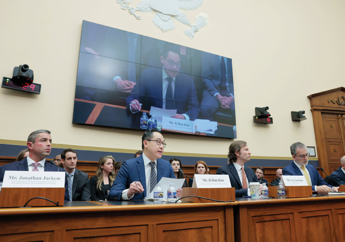 A man speaks at a congressional hearing, sitting at a long wooden table with nameplates and other officials under a large video screen.