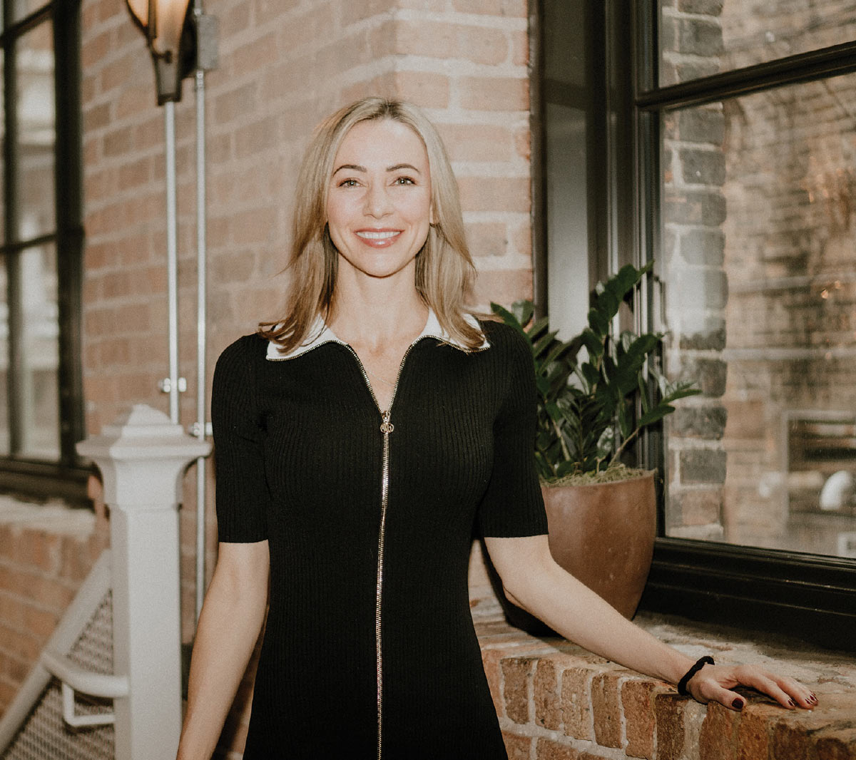A smiling woman in a black zip-up dress stands in an indoor space with exposed brick walls and large windows.