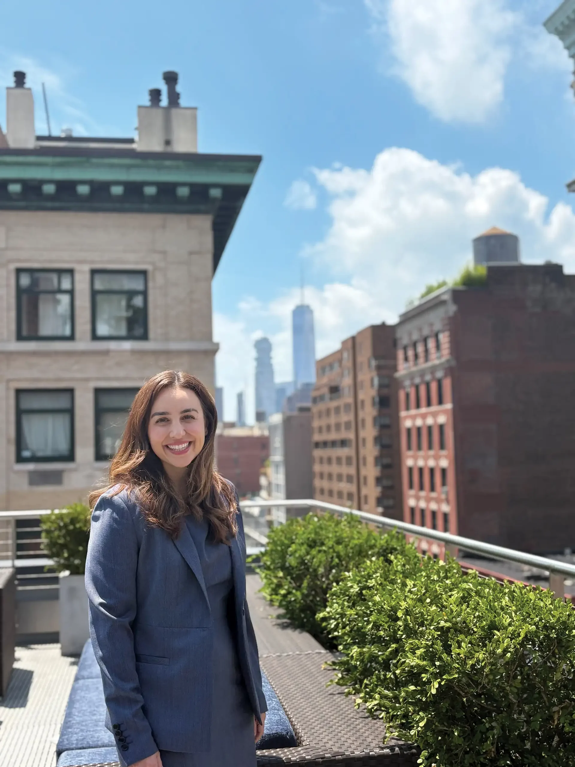 A smiling woman in a blue suit stands on a rooftop terrace, with the New York City skyline and One World Trade Center in the background.