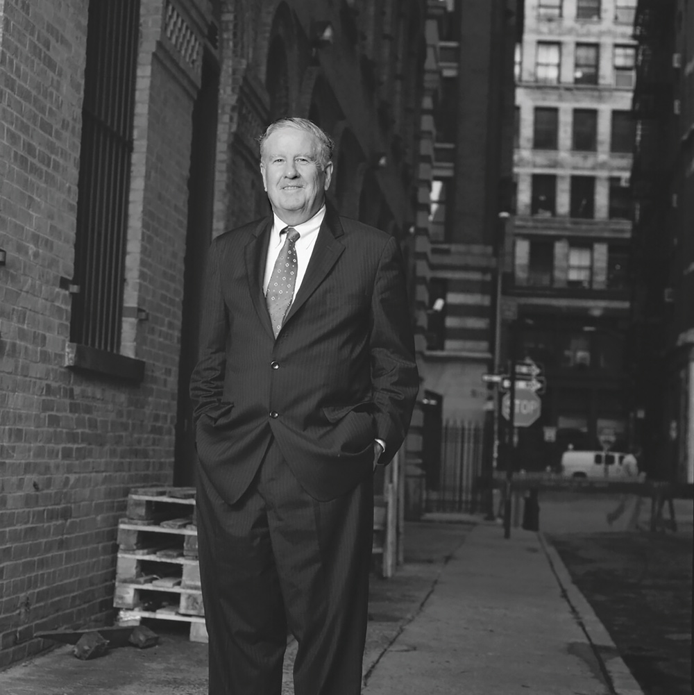 A black and white portrait of a man in a pinstriped suit standing in a city alleyway with his hands in his pockets.