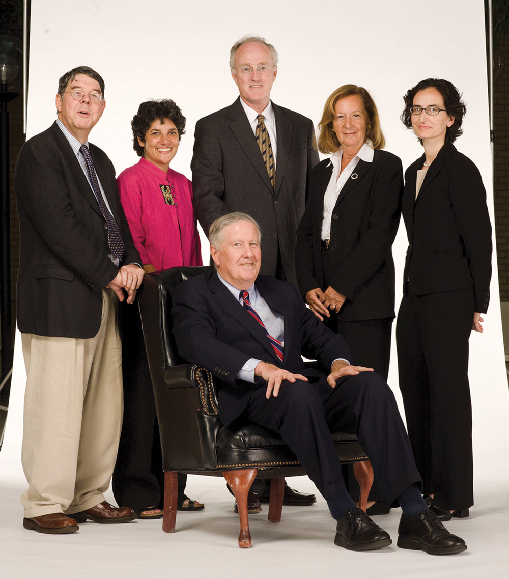 A man sits in a leather armchair surrounded by five standing colleagues, all in professional dress, on a white backdrop.