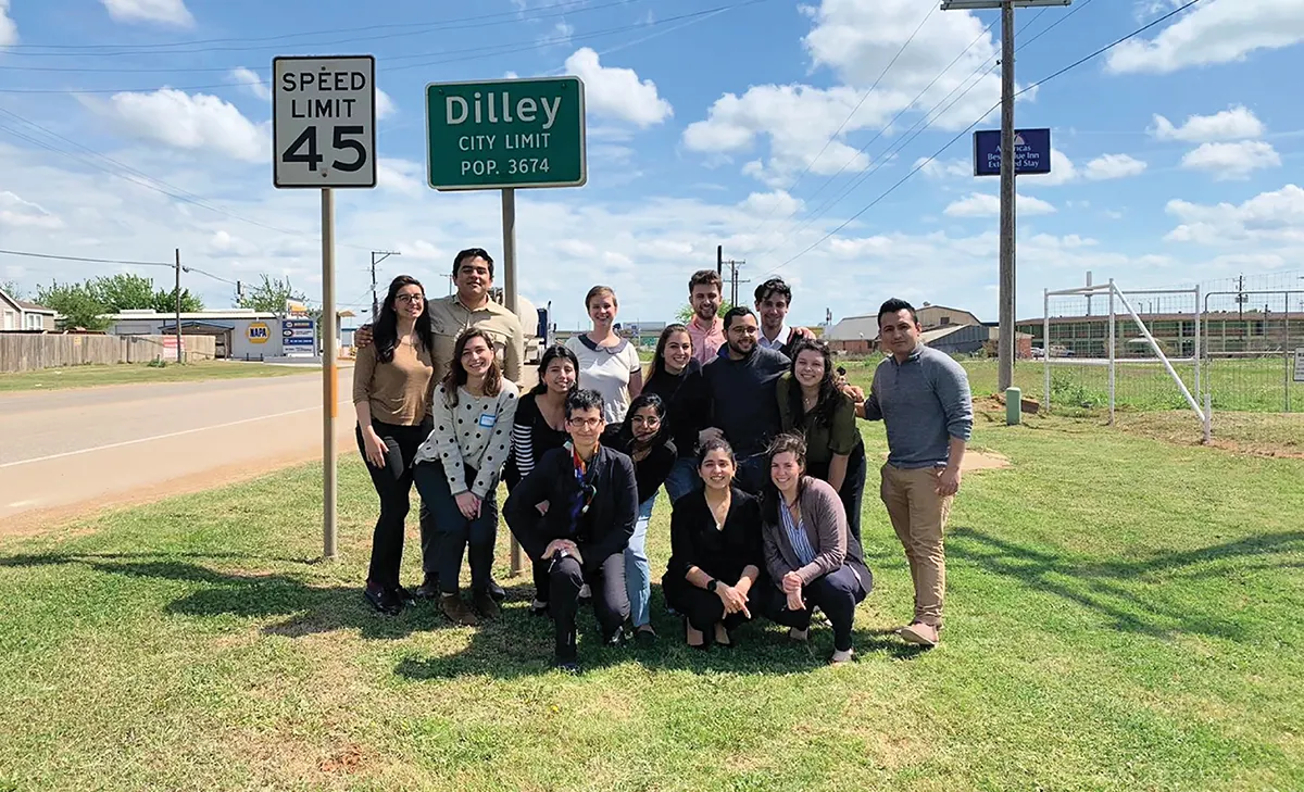 A large group of people posing for a photo on a grassy roadside next to a green "Dilley City Limit" sign.