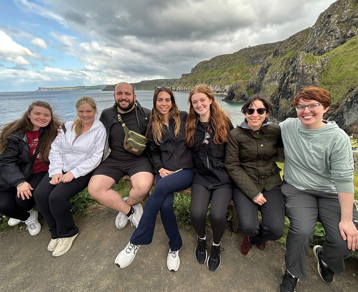 Seven young adults sitting on a bench along a coastal cliffside, smiling for a photo with a scenic backdrop of tall green cliffs and the ocean under a cloudy sky.