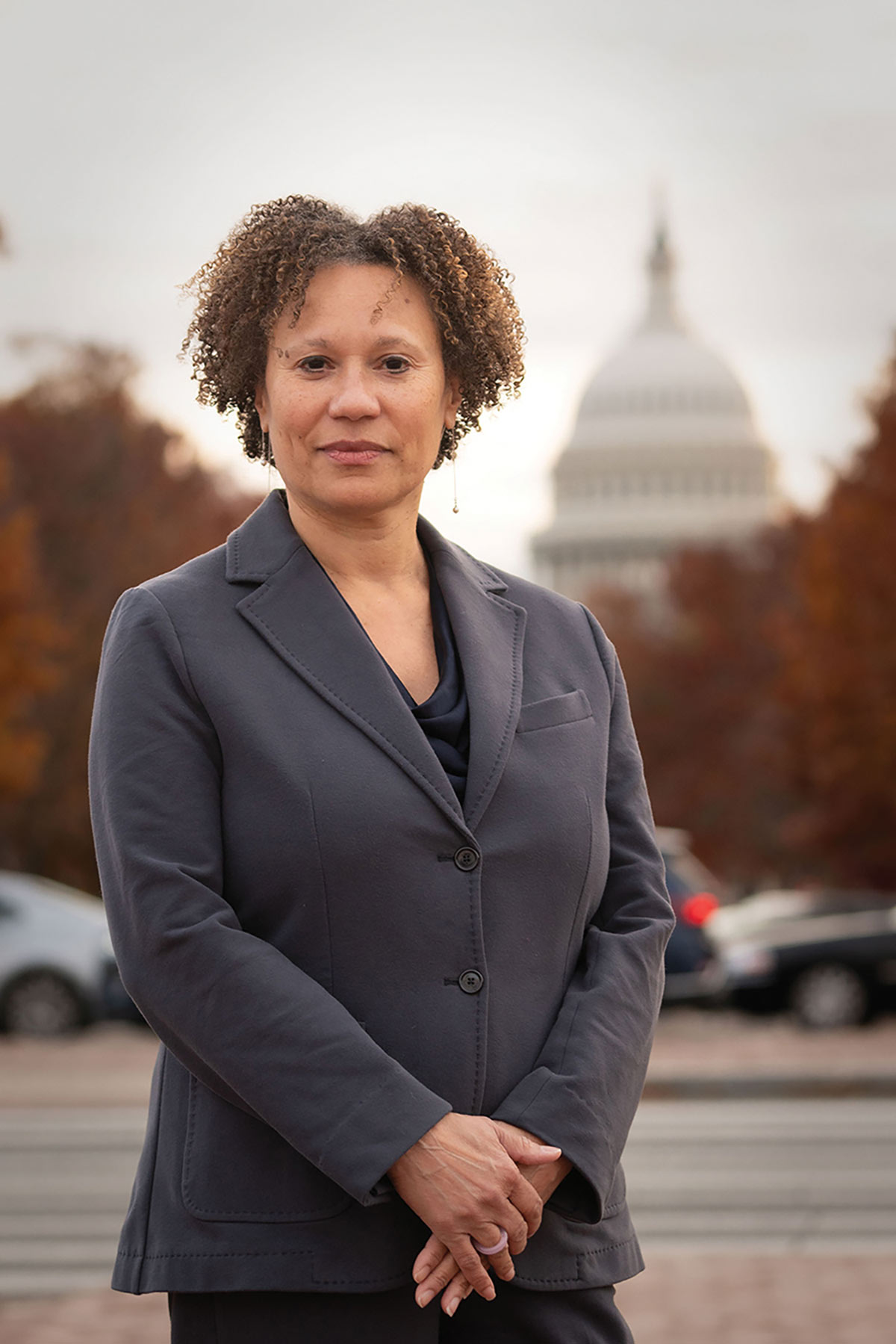 Catherine Powell in a grey blazer standing outdoors with the blurred U.S. Capitol dome in the background.