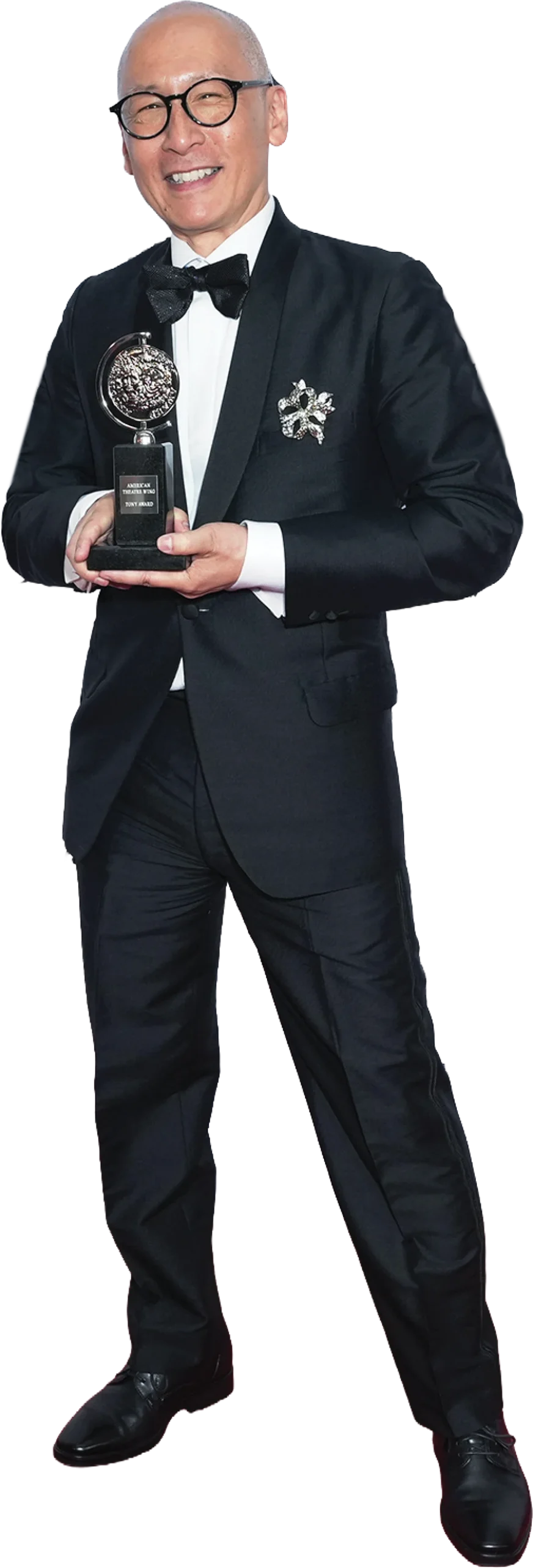 Smiling man in a black tuxedo and glasses holding a Tony Award. He features a sparkling brooch on his lapel and stands in a full-body pose against a white background.