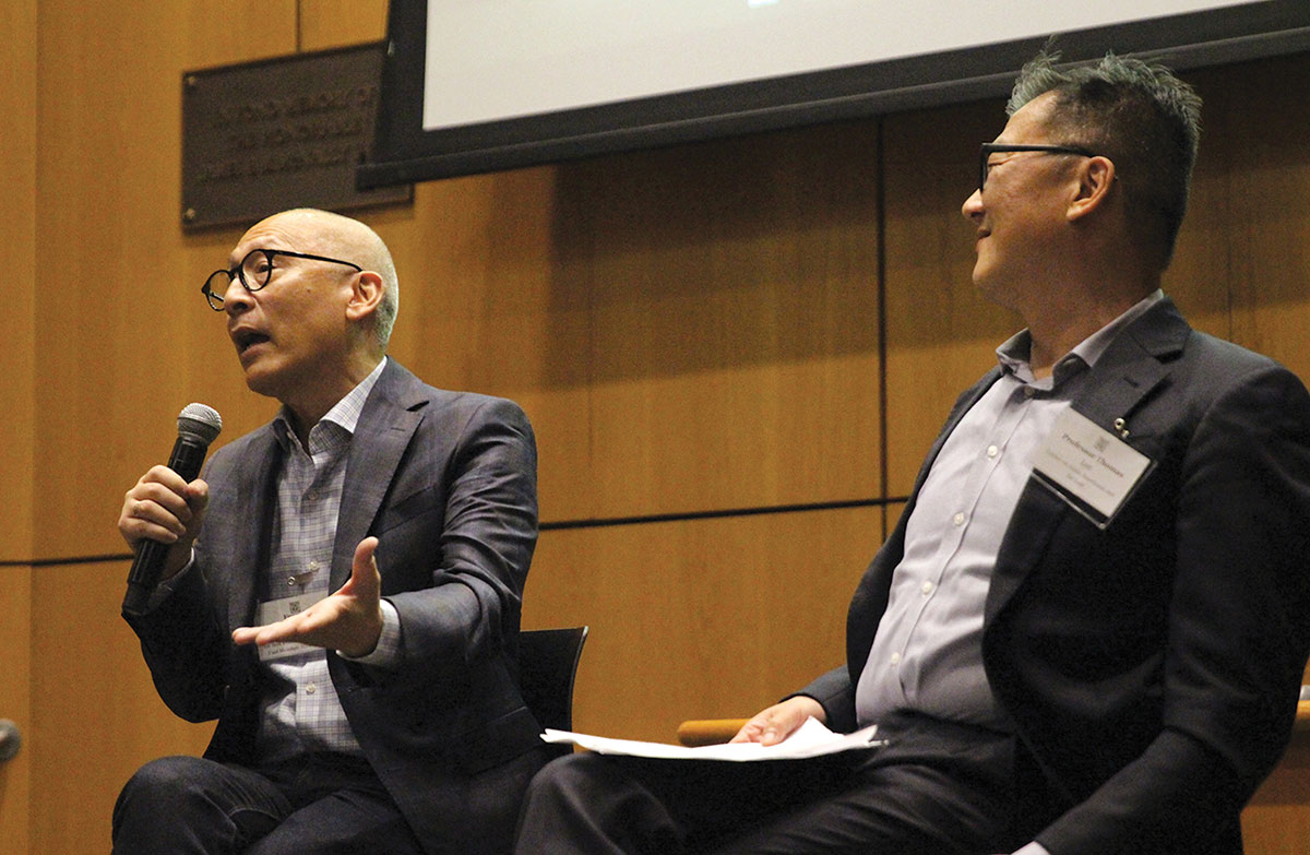 Two men in blazers participate in a panel discussion. The man on the left speaks into a microphone while gesturing, while Professor Thomas Lee sits to the right, smiling and listening against a wood-paneled wall.