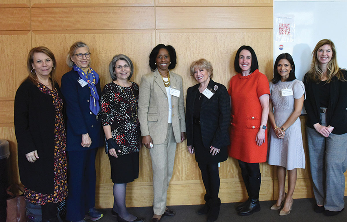 Eight women in professional attire pose for a group photo against a light wood-paneled wall.