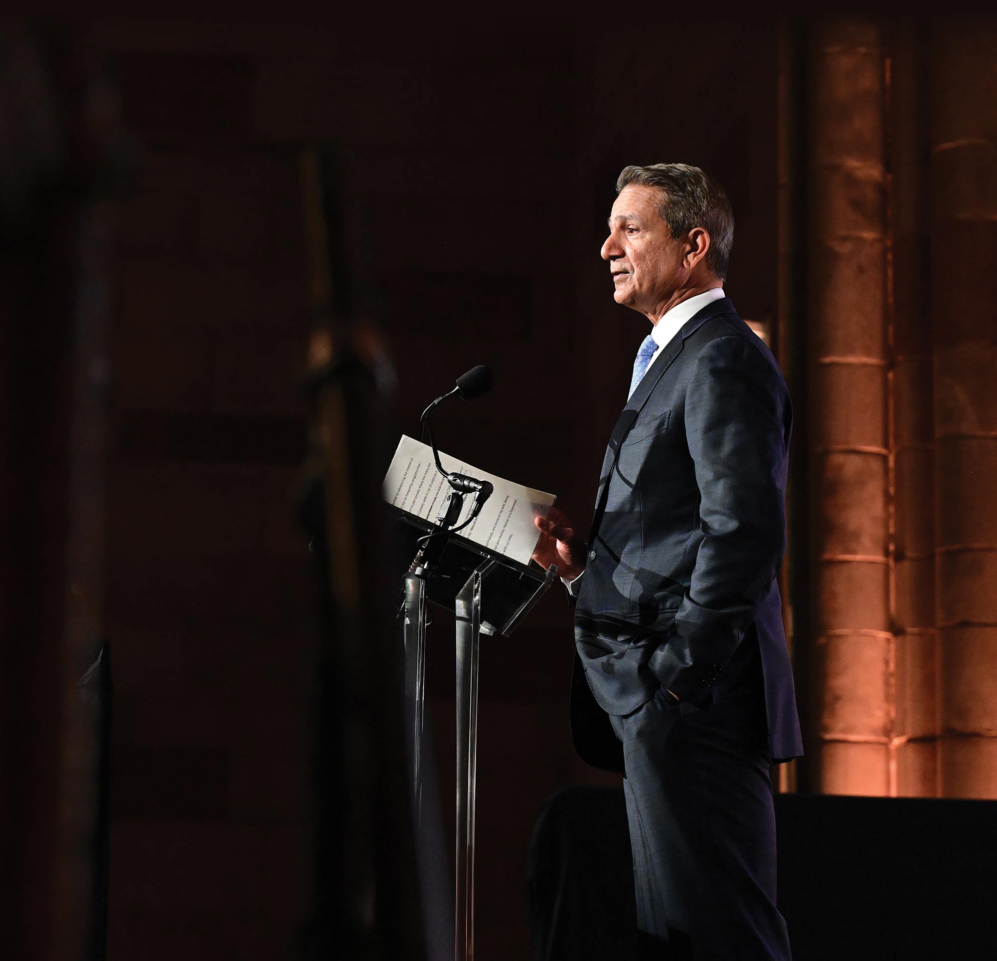 Sam Khichi in a dark suit and blue tie stands at a lectern with a microphone, holding notes while speaking at a formal event in a dimly lit, stone-walled hall.