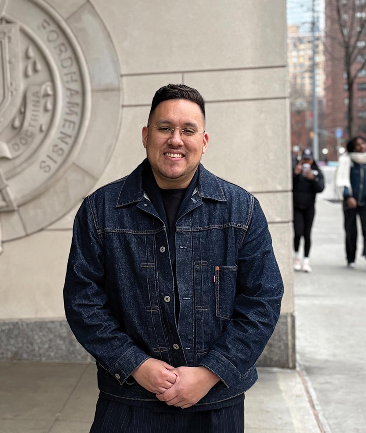 Christian Veliz wearing glasses and a denim jacket, standing in front of the Fordham University seal on a stone building.