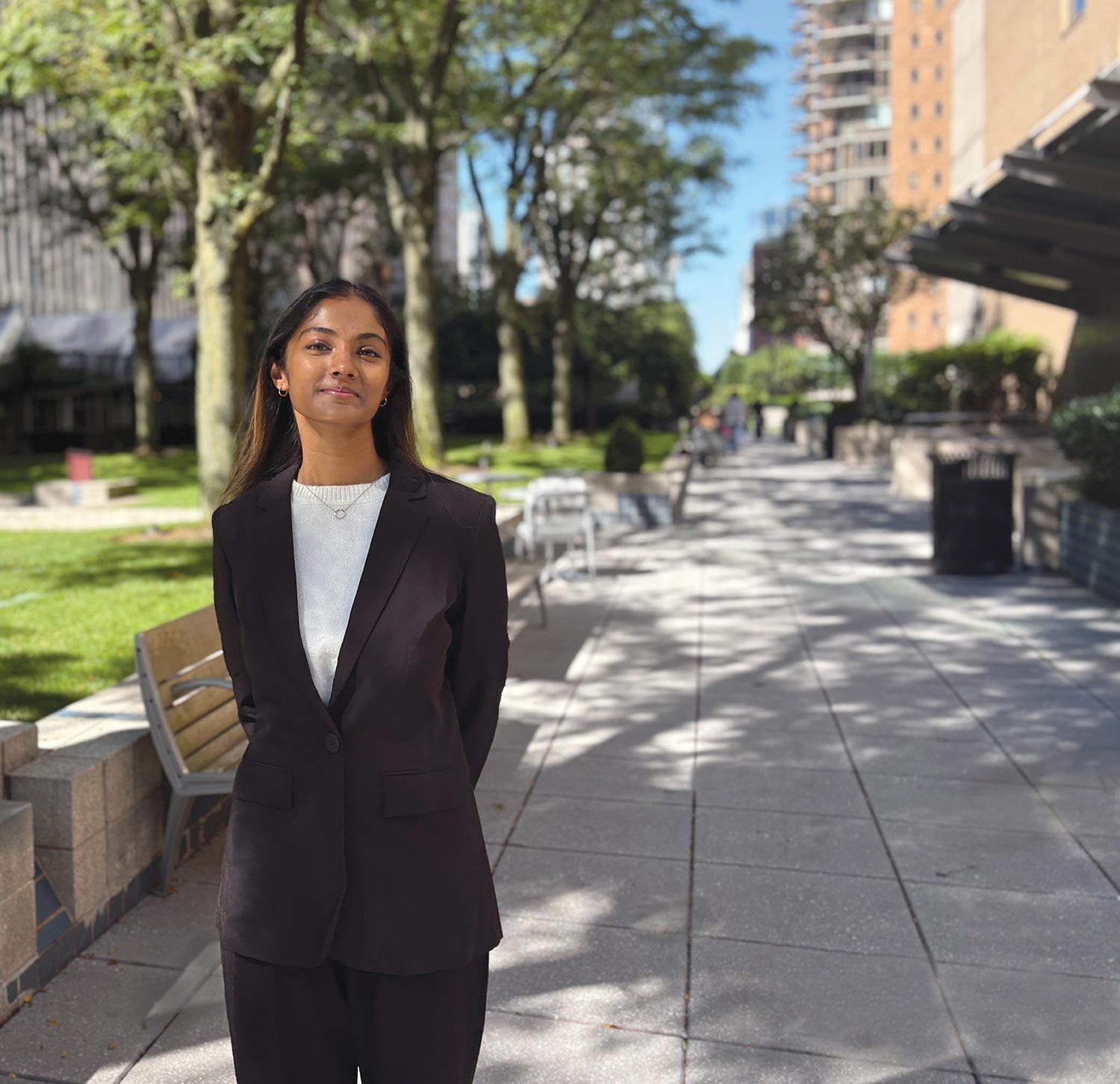 Amru Palaniyappan in a dark brown suit posing confidently on a sunny outdoor city walkway lined with trees and buildings.