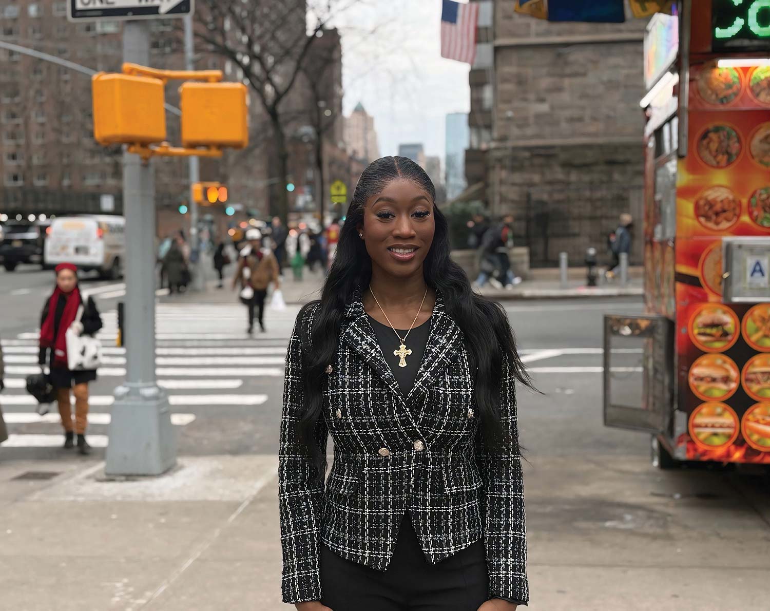 Adetoun Adelana in a black and white tweed blazer and gold cross necklace standing on a New York City street corner with a crosswalk and colorful food cart in the background.