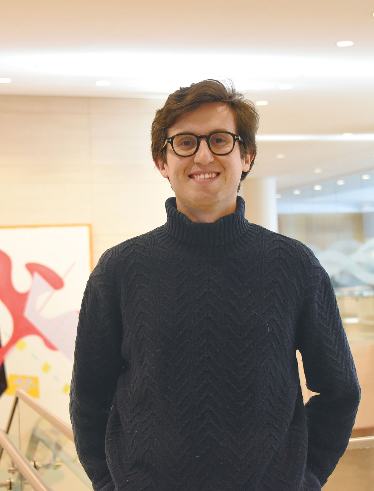 Juan Lozano de Diego wearing glasses and a navy blue cable-knit turtleneck sweater standing in a modern indoor setting with abstract art in the background.