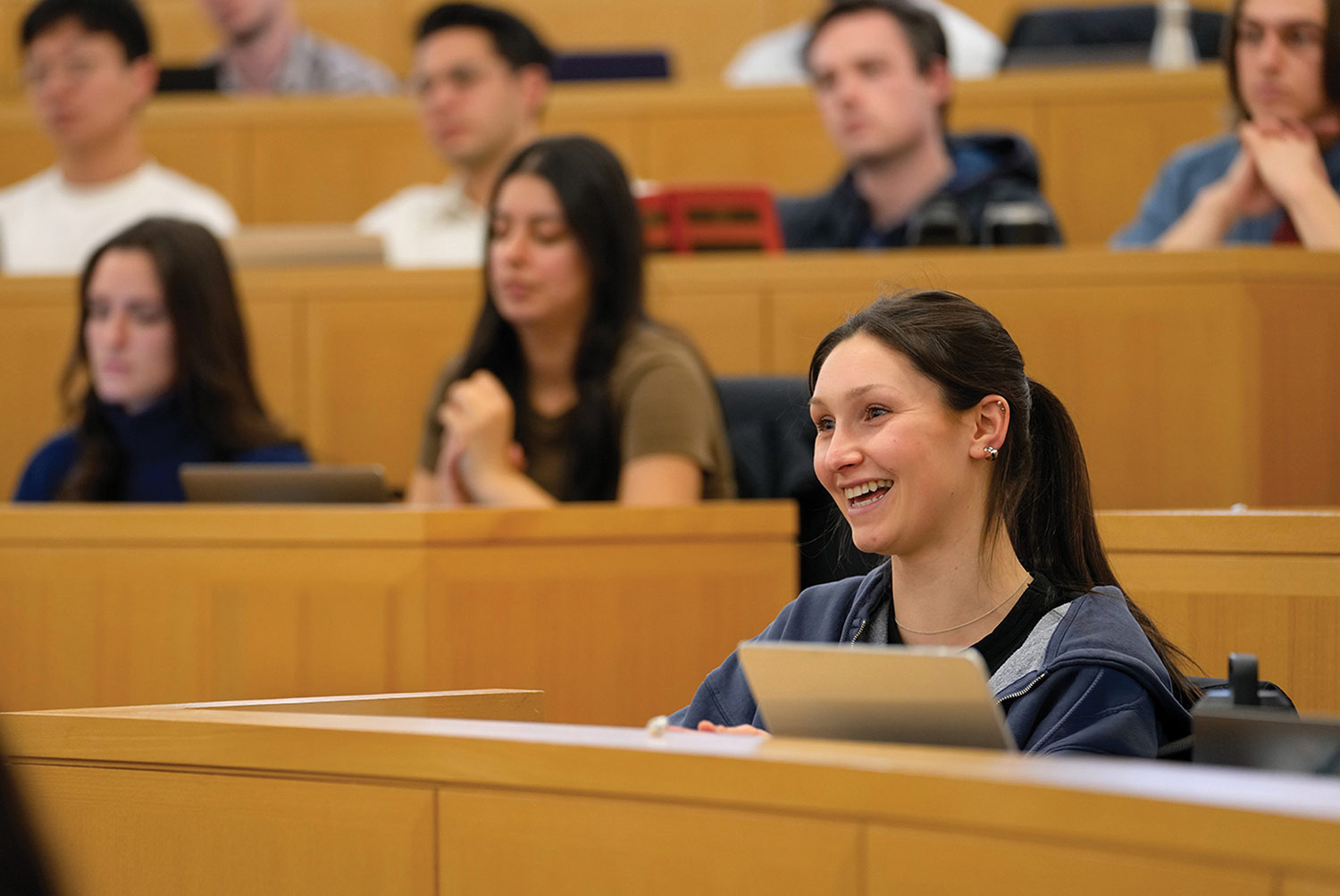 Smiling female student sits at a wooden desk in a tiered lecture hall with other students visible in the background.