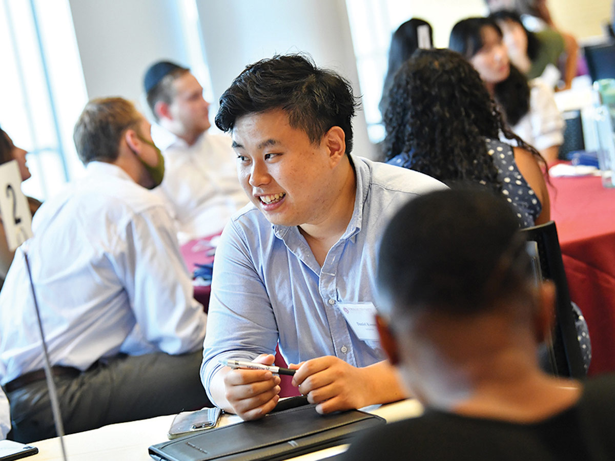 A smiling young man in a light blue button-down shirt talks to another attendee at a Fordham Law event, with red-clothed tables and other guests in the background.
