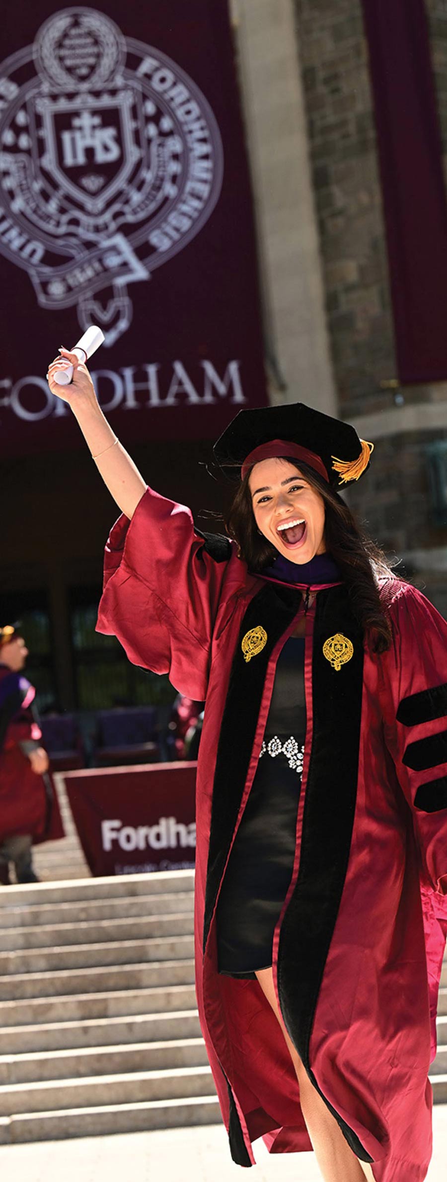 A joyful graduate in a maroon and black doctoral gown holds her diploma high in front of a Fordham University banner.