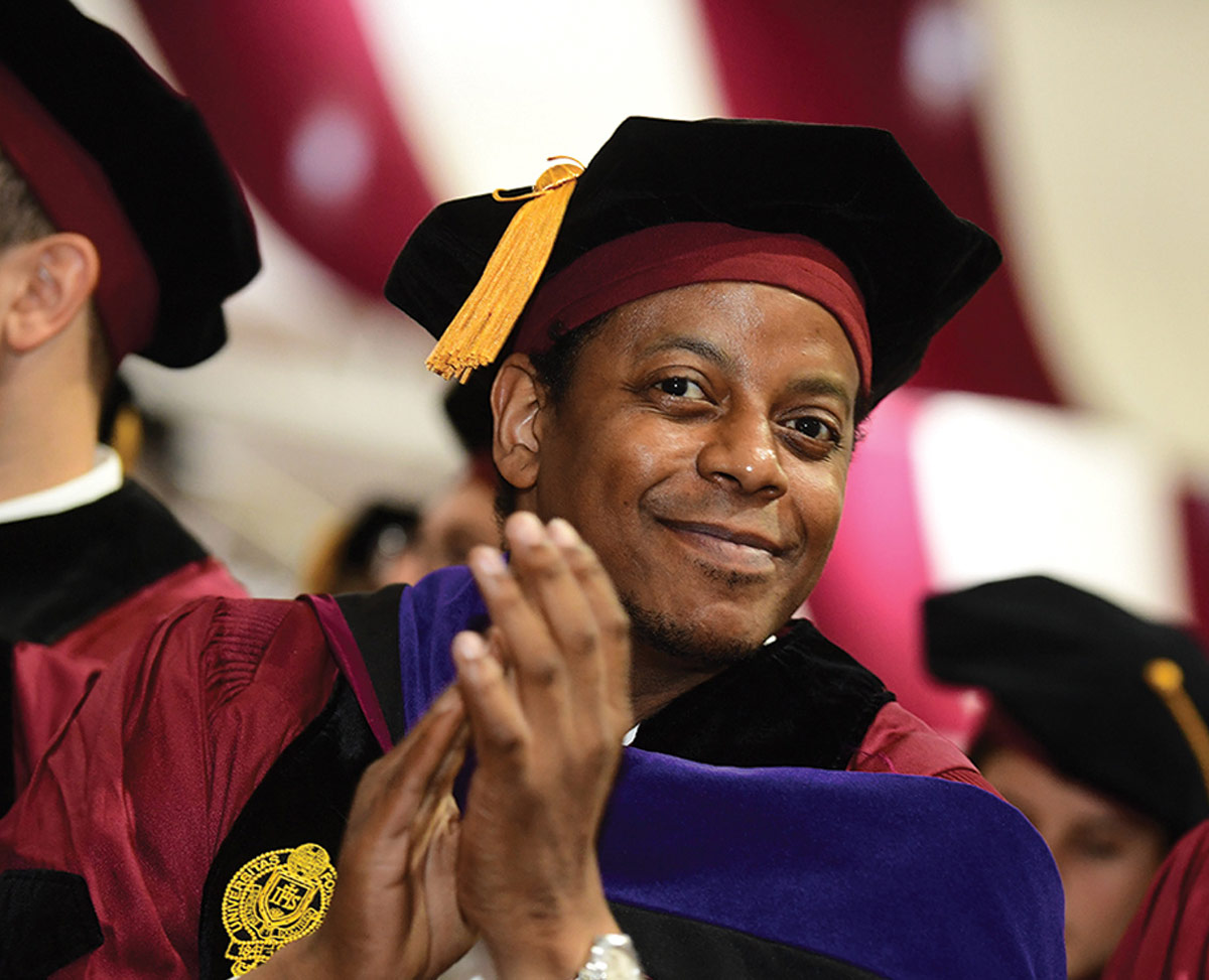 Smiling man in a maroon and black doctoral gown with a purple hood and gold-tasseled tam claps during a Fordham graduation ceremony.