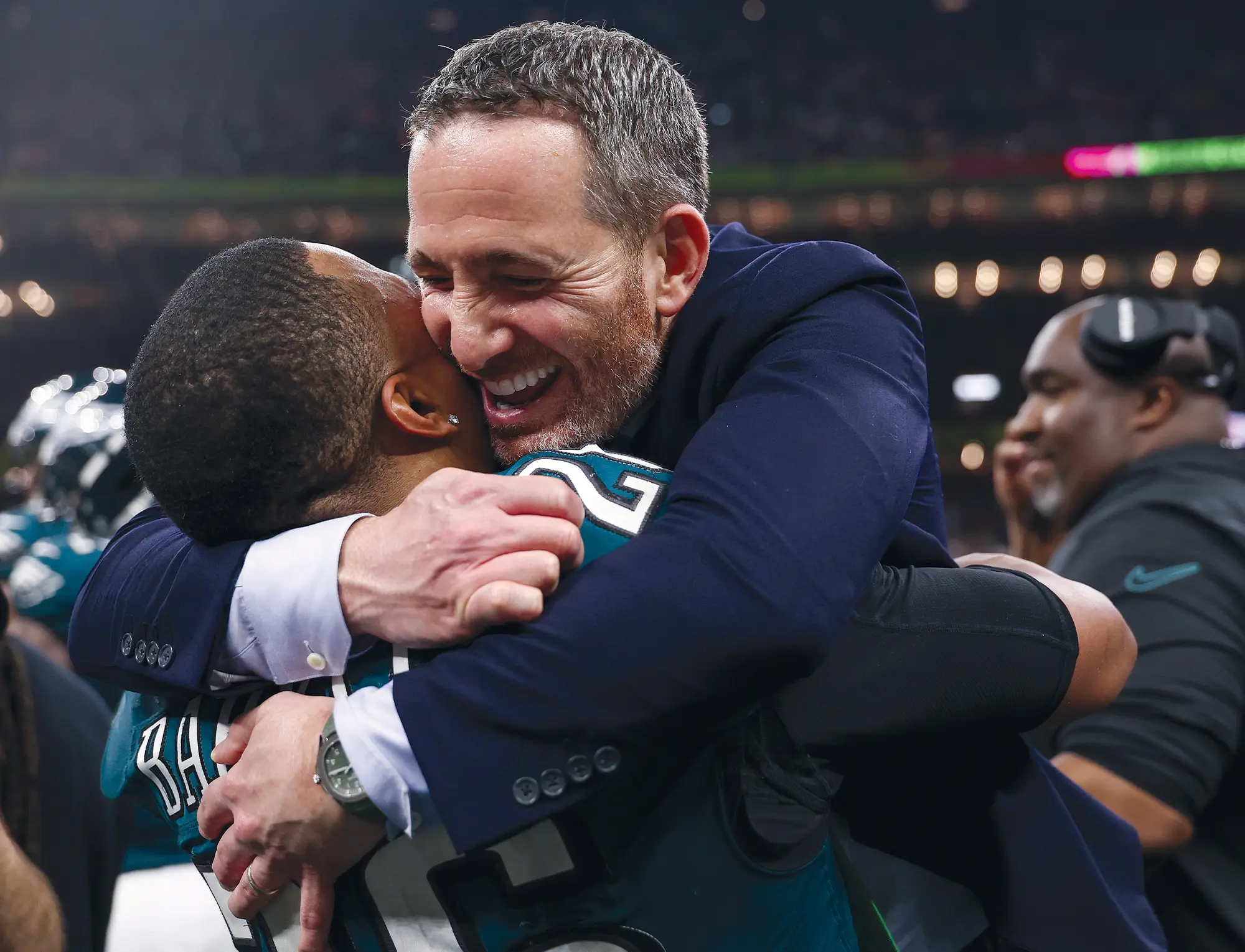 Howie Roseman, wearing a suit, smiles while hugging Saquon Barkley on a football field. Barkley wears a green Philadelphia Eagles jersey with the number 26 visible on the shoulder. Stadium lights are blurred in the background.