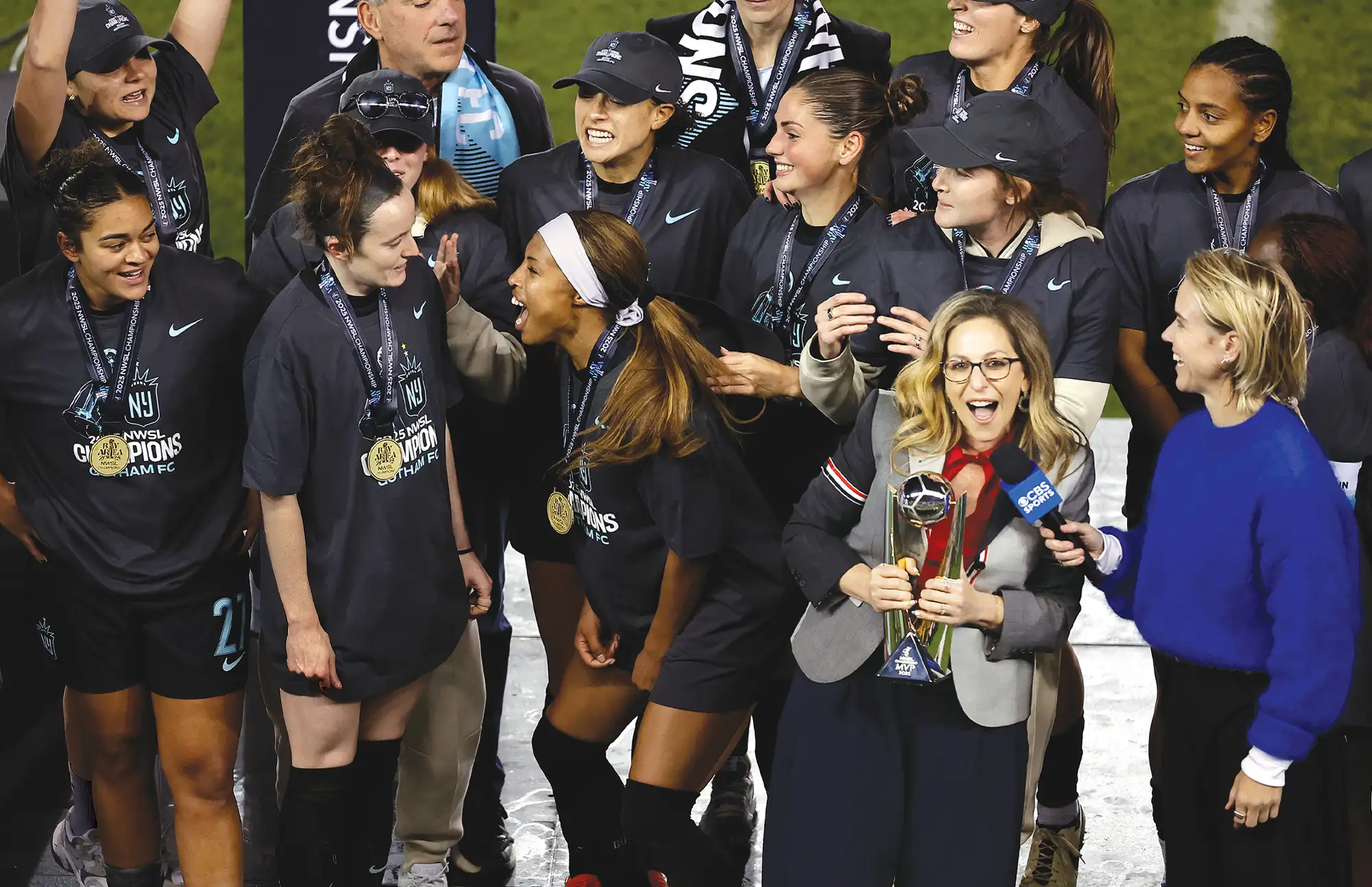 Jessica Berman holds a silver trophy and speaks into a CBS Sports microphone. Behind her, a group of soccer players wearing "NWSL Champions" t-shirts and gold medals celebrate on the field.