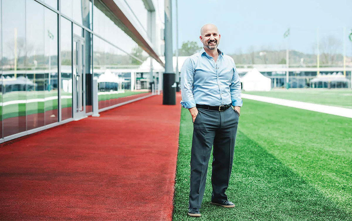 Andrew Lee stands with hands in his pockets on a green turf field next to a red path. He wears a light blue button-down shirt and grey slacks. A glass building on the left reflects the field and surrounding athletic facility.