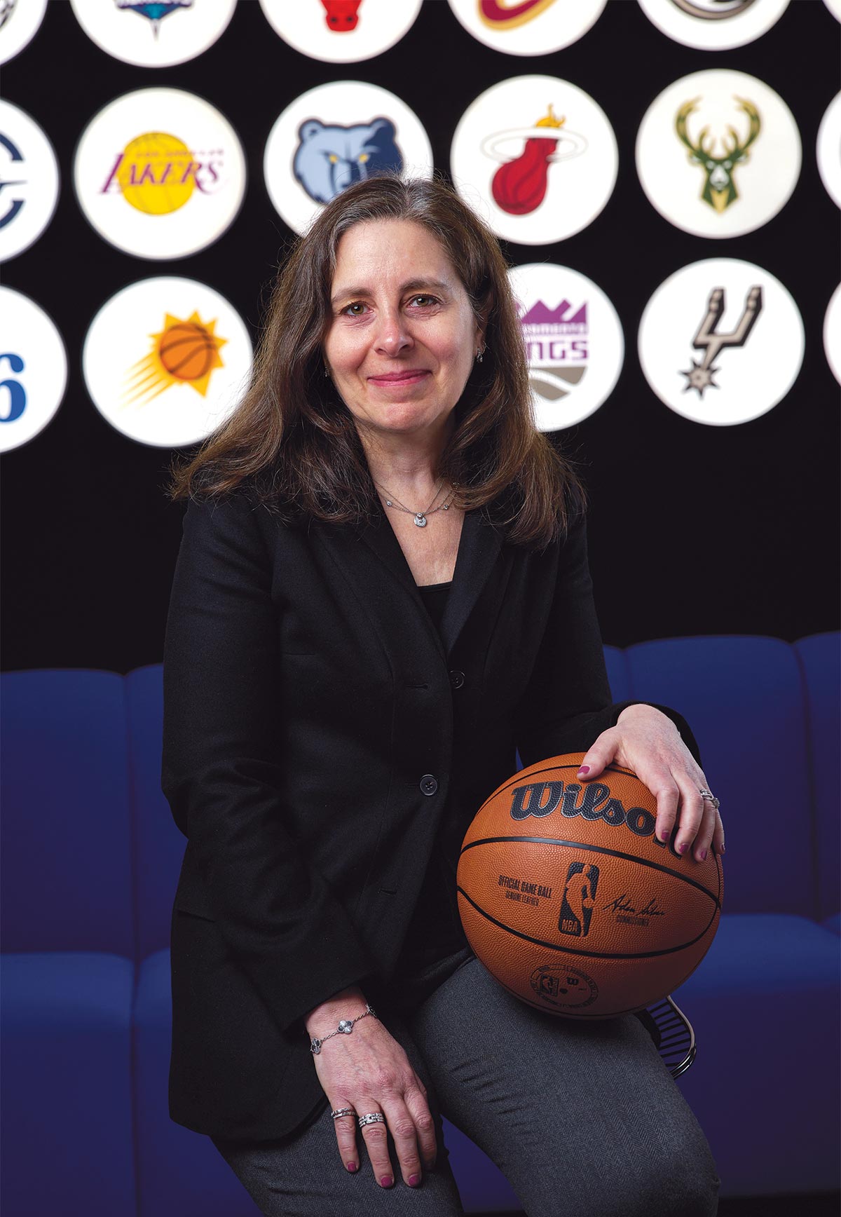 Elizabeth Maringer sits wearing a black blazer and rests her hand on an NBA basketball. The background features a series of circular, illuminated NBA team logos against a black wall.