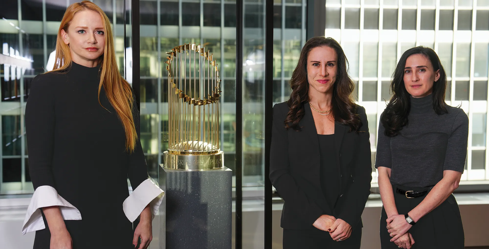 Marianne Adams, Jessica Saks, and Julia Vogel stand in an office setting with the gold Commissioner’s Trophy on a pedestal. Large windows behind them overlook a city building.