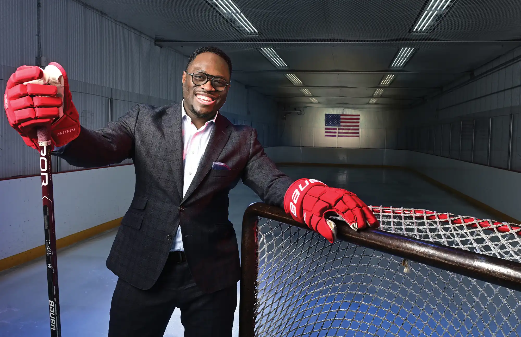 Osei-Tutu stands on an ice rink wearing a suit and red hockey gloves. He holds a hockey stick upright in his right hand and leans his left arm on a hockey net. An American flag is mounted on the wall in the background.