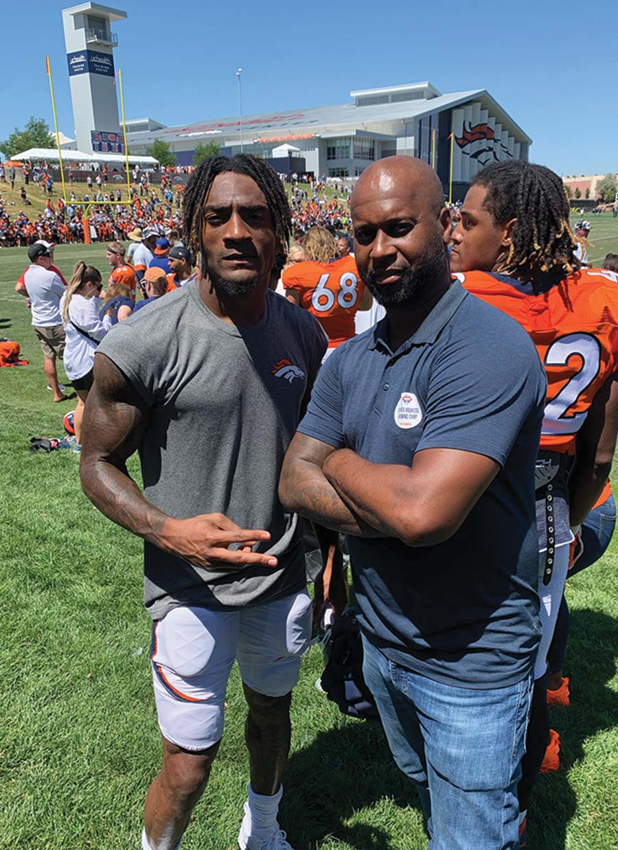 Ford stands with arms crossed next to Blessaun Austin on a practice field. Austin wears a grey Broncos practice shirt. A large athletic training facility and stadium tower are visible in the background.