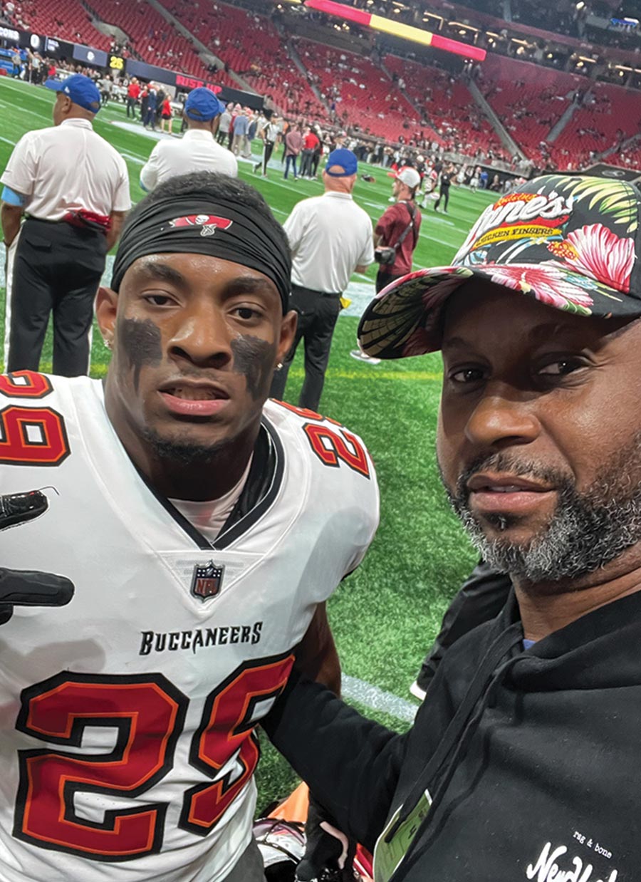Ford, wearing a floral hat, takes a selfie with Christian Izien. Izien wears a white Tampa Bay Buccaneers jersey and black eye grease on a football field with red stadium seating in the background.