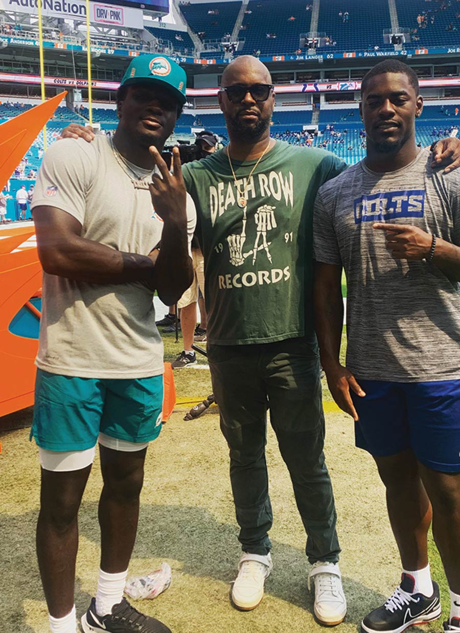Ford stands between Trill Williams and Deon Jackson on a football field. Williams wears a teal Dolphins hat and Jackson wears a grey Colts t-shirt. Ford wears a green "Death Row Records" shirt.